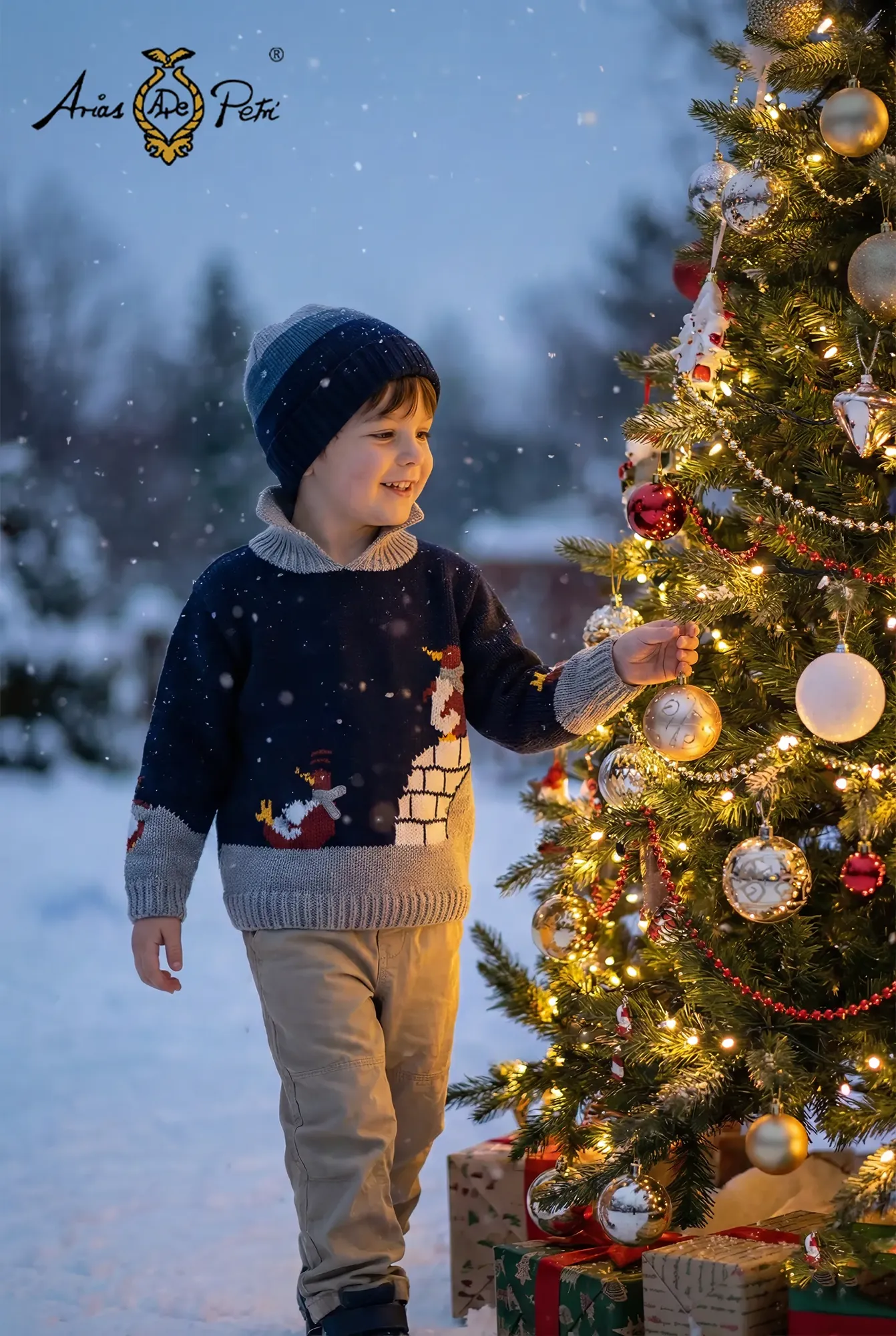 Lächelnder Junge mit gestreifter Mütze im dunkelblauen Strickpullover mit Pinguin-Motiv schmückt einen beleuchteten Weihnachtsbaum draußen im Schnee.